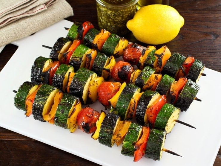 Four grilled and cooked lemon pesto vegetable skewers on white plate on wooden table, lemon, pesto and oil in background with napkin.