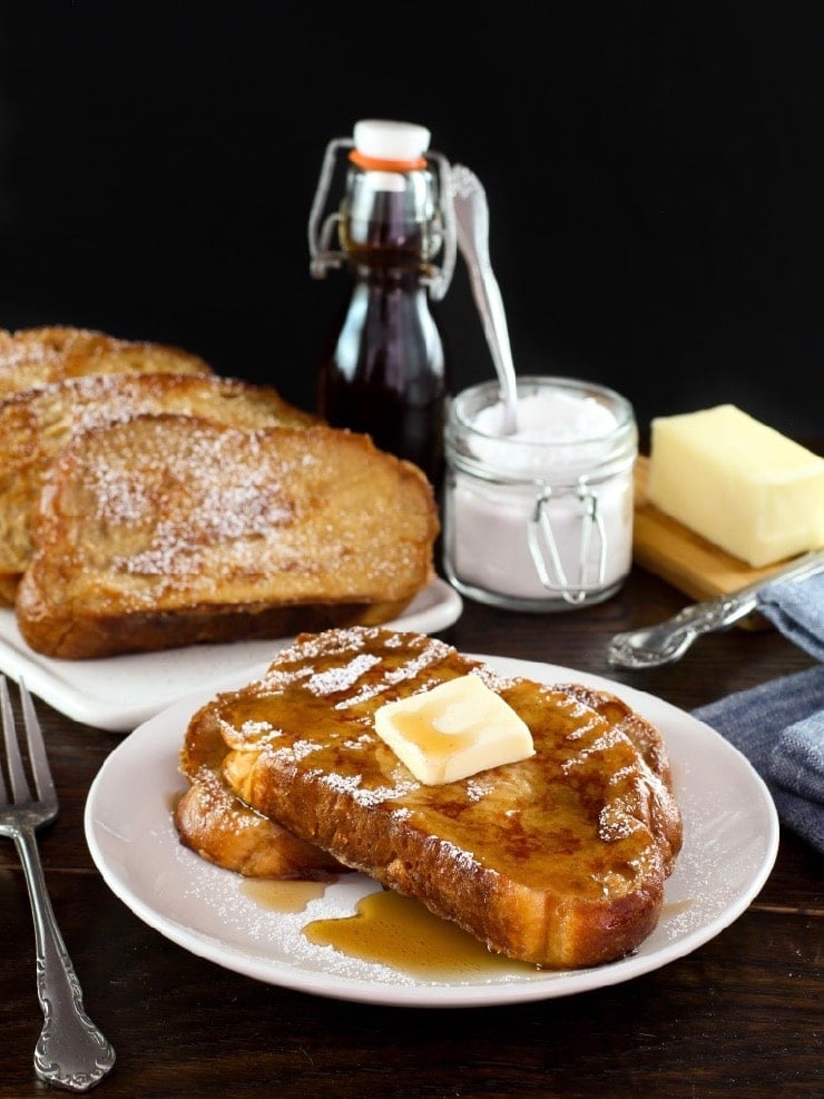 Vertical shot of Challah French Toast topped with pat of butter of maple syrup on a plate, platter of French toast, powdered sugar, bottle of maple syrup and stick of butter in background.