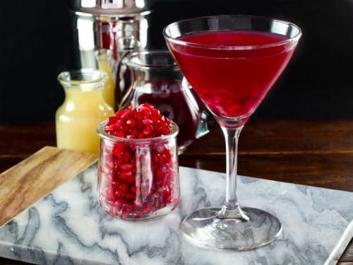 Martini glass with pomegranate martini on marble cutting board, glass carafe of pomegranate seeds, glass carafe of lime juice, martini shaker in background, on dark wooden surface.