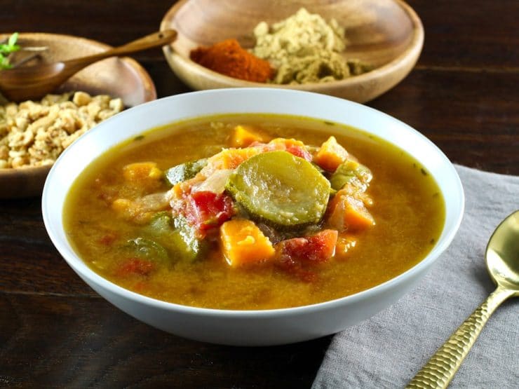 Autumn Sweet Potato Soup - whole vegetables in broth served in white bowl with napkin and spoon. Wooden bowls with fresh thyme, peanuts and spices in the background. Horizontal shot.