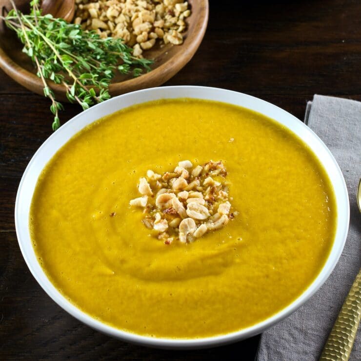 Square featured shot - Pureed Autumn Sweet Potato Soup with roasted peanut garnish in white bowl with napkin and spoon. Wooden bowls with fresh thyme, peanuts and spices in the background.