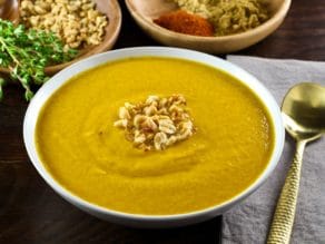 Pureed Autumn Sweet Potato Soup with roasted peanut garnish in white bowl with napkin and spoon. Wooden bowls with fresh thyme, peanuts and spices in the background. Horizontal shot.