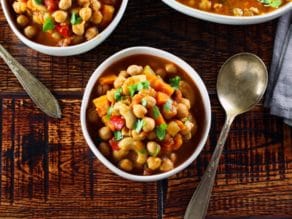 Overhead shot of chickpea chili garnished with cilantro with spoon on wooden table, two other bowls of chili on periphery.