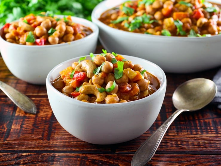 Frontal shot of white bowl of slow cooker vegan chickpea chili with spoon on table, two other bowls of chili and cilantro in background.