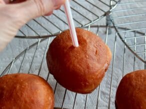 Sufganiyot with straw being inserted into the center by hand, on cooling rack.