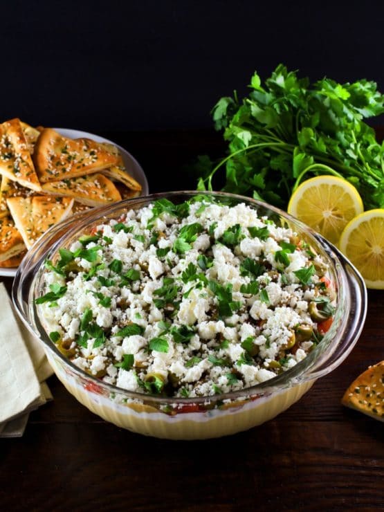 A large glass serving bowl filled with a layered dip that includes hummus, greek yogurt, Israeli salad, olives, feta cheese, and cilantro. A dish of pita chips is sitting off to the side.