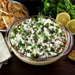 A large glass serving bowl filled with a layered dip that includes hummus, greek yogurt, Israeli salad, olives, feta cheese, and cilantro. A dish of pita chips is sitting off to the side.