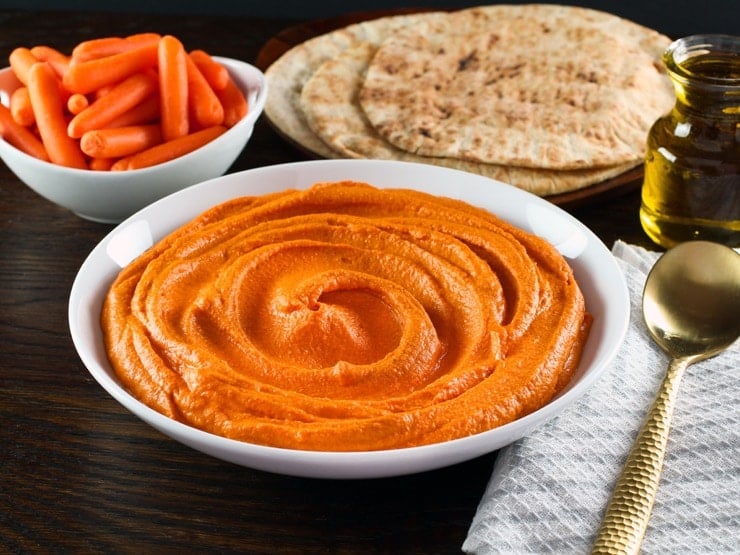 Horizontal shot - white bowl of red pepper hummus with gold spoon, cloth napkin on side. Pita and dish of carrots in background.