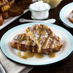 Wider horizontal shot - plate of challah french toast drizzled with Kahlua cream sauce, napkin and platter of french toast in background, powdered sugar with small spoon behind.