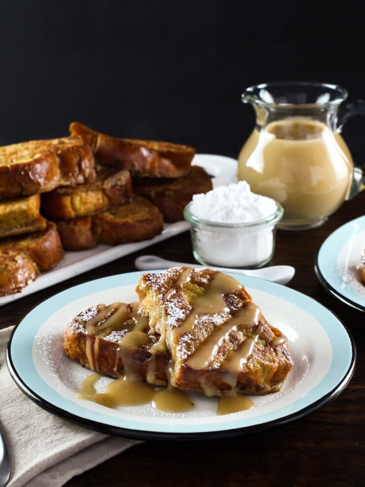 Vertical shot - plate of 2 slices of challah french toast drizzled with Kahlua cream sauce, napkin and platter of french toast in background, powdered sugar with small spoon and carafe of cream sauce behind.