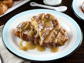 Close up horizontal shot - plate of challah french toast drizzled with Kahlua cream sauce, napkin and platter of french toast in background, powdered sugar with small spoon behind.