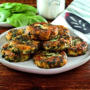 Horizontal shot - plate of fried keftes de espinaca - spinach keftes - piled on a white plate with fresh spinach, canister of breadcrumbs, spatula and cloth napkin in background.