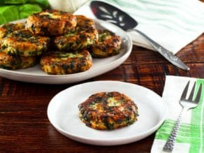 Horizontal shot - single fried spinach patty - on a white plate with fork and cloth napkin, plate of keftes piled on a white plate with spatula and cloth napkin in background.