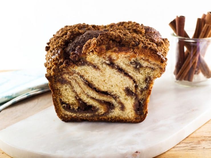 Babka loaf sliced, swirly cinnamon filling, on white marble cutting board with glass carafe of cinnamon sticks and cloth napkin in background.