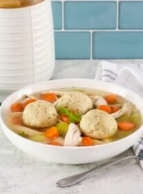 Matzo ball soup - horizontal shot of classic Jewish chicken soup with fluffy matzo balls on a white countertop. Spoon and linen napkin beside the bowl, fresh green herbs and white ceramic jar in background.