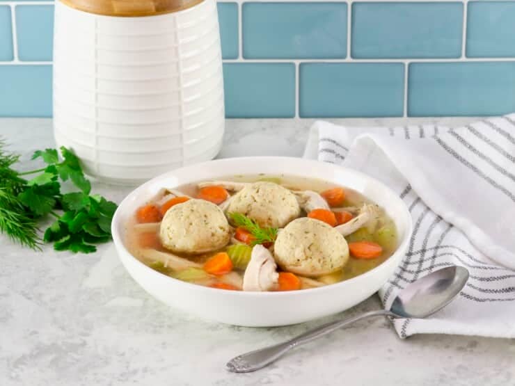 Matzo ball soup - horizontal shot of classic Jewish chicken soup with fluffy matzo balls on a white countertop. Spoon and linen napkin beside the bowl, fresh green herbs and white ceramic jar in background.