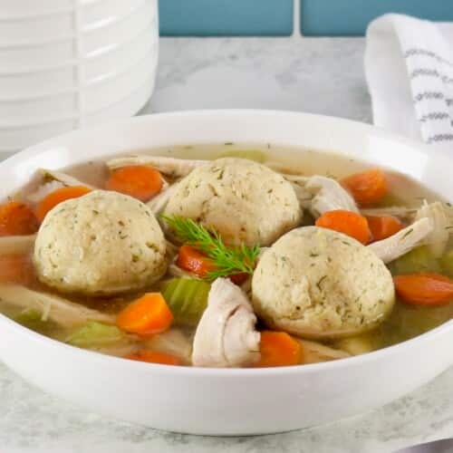 Horizontal shot of floater matzo balls in a shallow bowl of Jewish chicken soup with carrot slices, pieces of celery, and golden broth. Spoon and linen napkin on the white marble counter beside the bowl. Tiles and white jar in background.