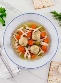 Overhead shot - sinker matzo ball soup on a white marble surface. Soup includes dense sinker matzo balls, chicken, carrots, celery, and golden broth in a white soup bowl with spoon, cloth napkin, cracked matzo and fresh green herbs beside the bowl. Classic Jewish soup.