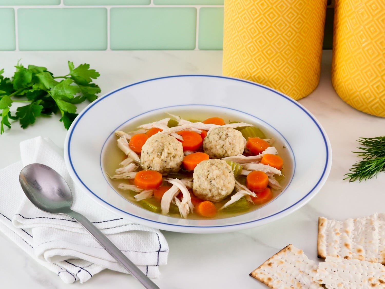 Floater matzo balls in a shallow bowl of Jewish chicken soup with carrot slices, pieces of celery, and golden broth. A spoon, fresh herbs, three small sheets matzo and two yellow large canisters in background.