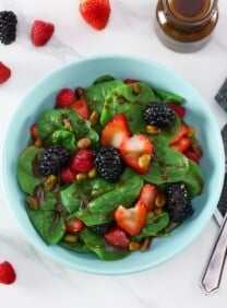 Overhead shot - spinach strawberry salad on a light blue plate topped with maple balsamic dressing, strawberries, blackberries, raspberries and pistachios in foreground. Table set with fork and gray and white stripe linen napkin. Garnish of four small strawberries and two blackberries, and a small glass carafe bottle with maple balsamic dressing, in background.