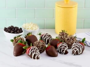 Chocolate dipped strawberries, some with nuts and some chocolate strawberries drizzled with white chocolate in foreground, a yellow canister, linen napkin and two small bowls with white and dark chocolate chips separately in background.