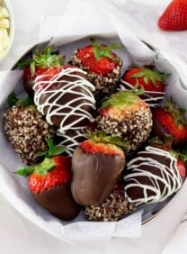 Chocolate dipped strawberries, some with nuts and some chocolate strawberries drizzled with white chocolate on a parchment lining paper inside a medium-size circular gold tin can in foreground, with a linen napkin on the side. Three fresh strawberries, and two small bowls with white and dark chocolate chips separately in background.