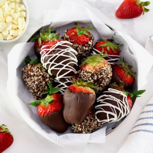 Chocolate dipped strawberries, some with nuts and some chocolate strawberries drizzled with white chocolate on a parchment lining paper inside a medium-size circular gold tin can in foreground, with a linen napkin on the side. Three fresh strawberries, and two small bowls with white and dark chocolate chips separately in background.