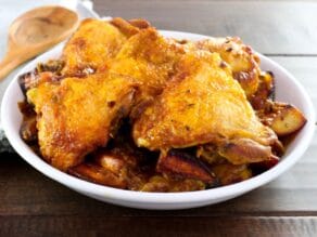 Israeli Chicken Sofrito - braised chicken pieces in a large shallow white bowl on wooden table with wooden spoon and napkin in background.