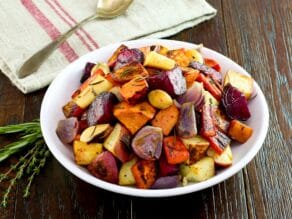 Horizontal shot - plate of roasted root vegetables, rosemary and thyme on the side, cloth napkin and spoon in background, on a wooden table.