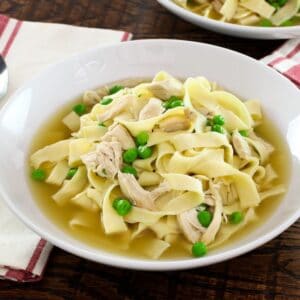 Horizontal shot - bowl of chicken with homemade noodles and sweet peas with cloth napkin and spoon on wooden background.