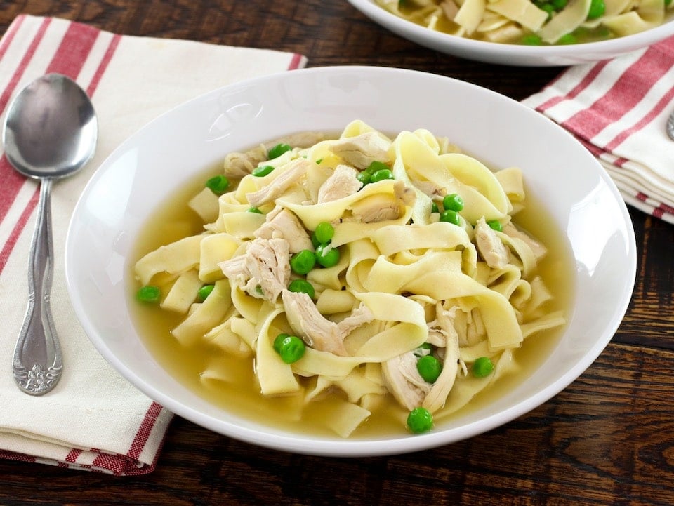 Horizontal shot - bowl of chicken with homemade noodles and sweet peas with cloth napkin and spoon on wooden background.