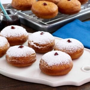 Sufganiyot on white platter dusted with sugar, more in background on cooling rack, with blue cloth towel.