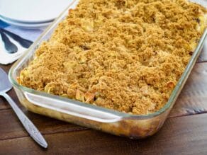 A glass dish of noodle kugel with graham cracker topping on wooden table, utensil and napkin beside it.