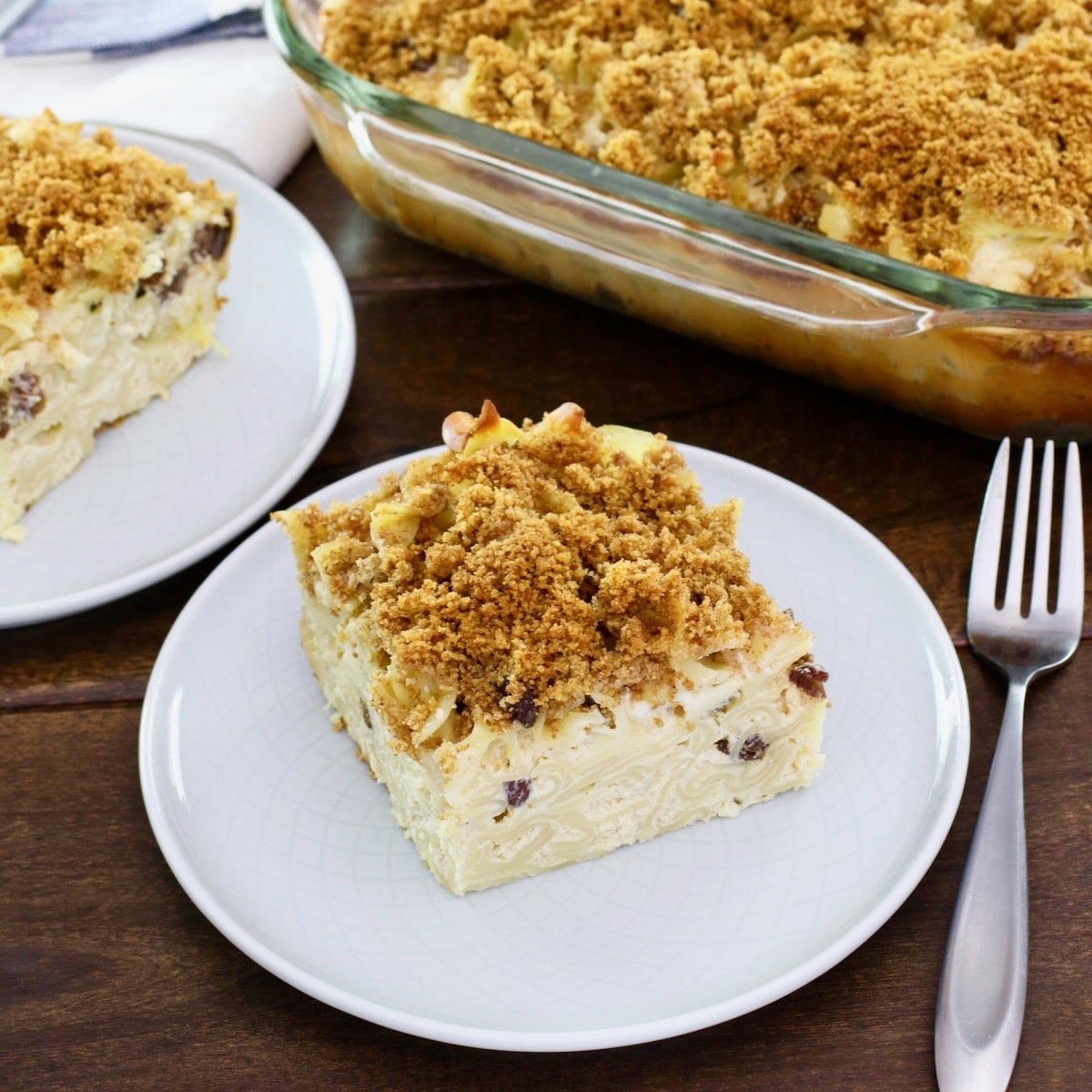 Square Crop - a piece of kugel on a small plate with fork on wooden table, dish of kugel and another slice in background.