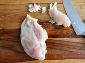 Chicken breast with tenderloin sliced off, knife and trimmings beside, on wooden cutting board.