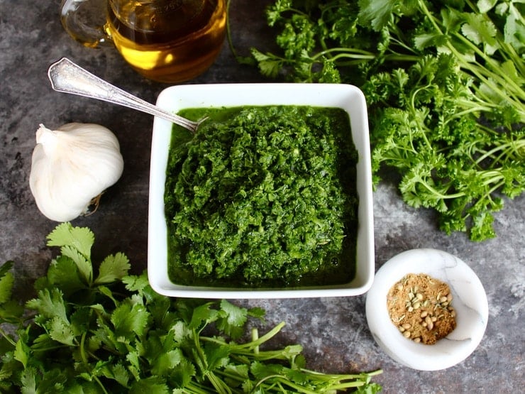 Overhead view of Yemenite Schug sauce in white square bowl with spoon surrounded by fresh herbs, head of garlic, olive oil in glass carafe and spices in white marble bowl on countertop