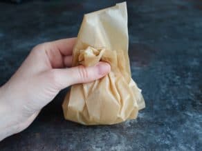 Hand gathering parchment around whole head of garlic on grey countertop.