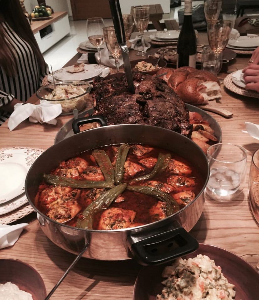 A Shabbat table set with pan of paprika pepper fish, meat, challah, and salad. Utensils, plates, and glasses for water. Wine bottle and half filled wine glasses in background. 