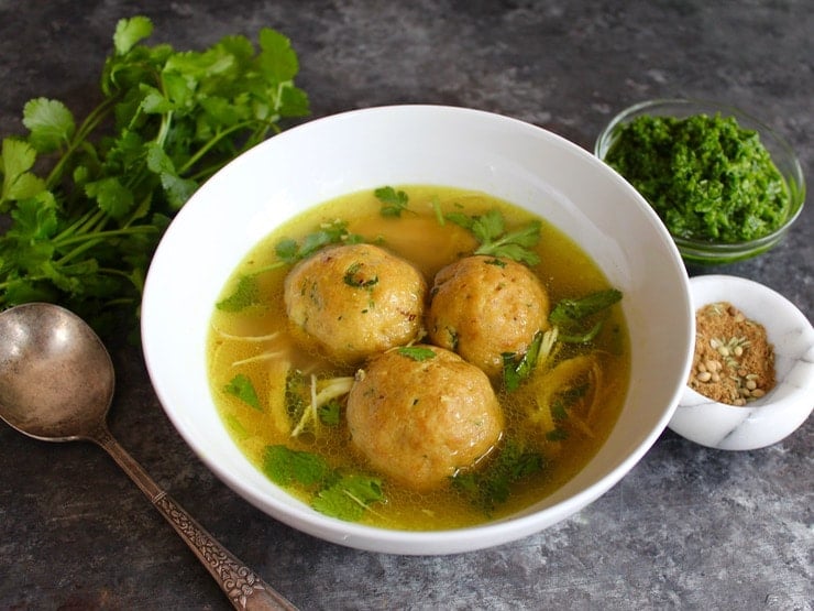 Yemenite matzo ball soup with three large matzo balls in a white bowl on a silver spoon surrounded with vegetables