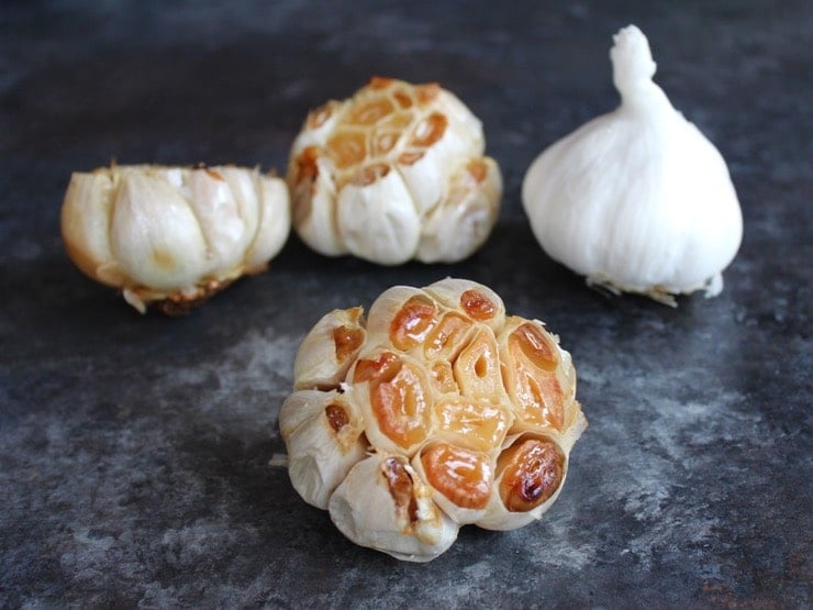 Head of roasted garlic with exposed cloves in foreground, three heads of garlic in background, one raw two roasted, on grey countertop.