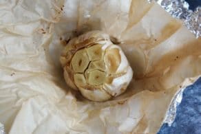 Garlic head with exposed cloves, roasted but not caramelized on layer of parchment on top of foil on grey countertop.