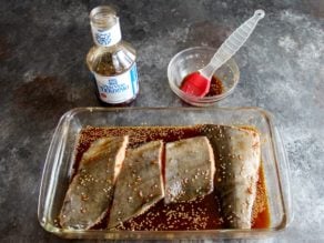 4 pieces of cod fillet in a 9x13 glass dish covered with Soy Vay Veri Veri Teriyaki Marinade and Sauce, bowl and brush with bottle of Soy Vay marinade in background.