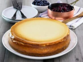 Rich smooth cheesecake baked golden on a plate, with blueberry topping and blueberries in background. Utensils and plates behind.