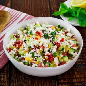 Horizontal shot - bowl filled with fresh Israeli Salad with Feta, tomatoes, cucumbers and bell peppers. Mint and lemon in the background, cloth napkin and wooden spoon beside the bowl.