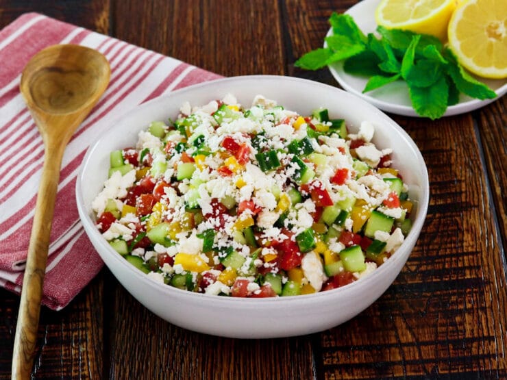 Horizontal shot - bowl filled with fresh Israeli Salad with Feta, tomatoes, cucumbers and bell peppers. Mint and lemon in the background, cloth napkin and wooden spoon beside the bowl.