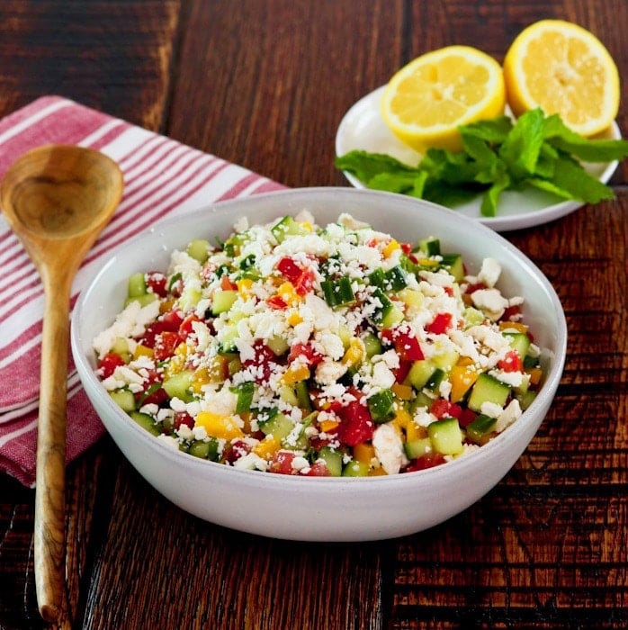 Square crop - bowl filled with fresh Israeli Salad with Feta, tomatoes, cucumbers and bell peppers. Mint and lemon in the background, cloth napkin and wooden spoon beside the bowl.