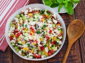 Vertical overhead shot - large white bowl filled with Israeli Salad with Feta Cheese, wooden serving spoon and cloth napkin beside, dish of fresh mint in background.