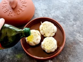 Bottle drizzling olive oil on one of three garlic heads with exposed cloves in ceramic garlic roaster on grey countertop, roaster lid in background.