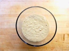 Sourdough starter in a glass jar bubbling on a wooden cutting board.