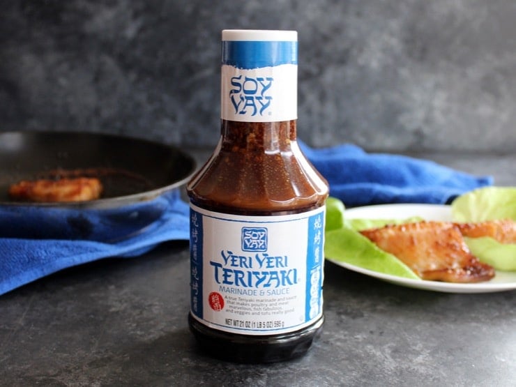 Bottle of Soy Vay Veri Veri Teriyaki Marinade and Sauce, fish and skillet in background with blue towel.